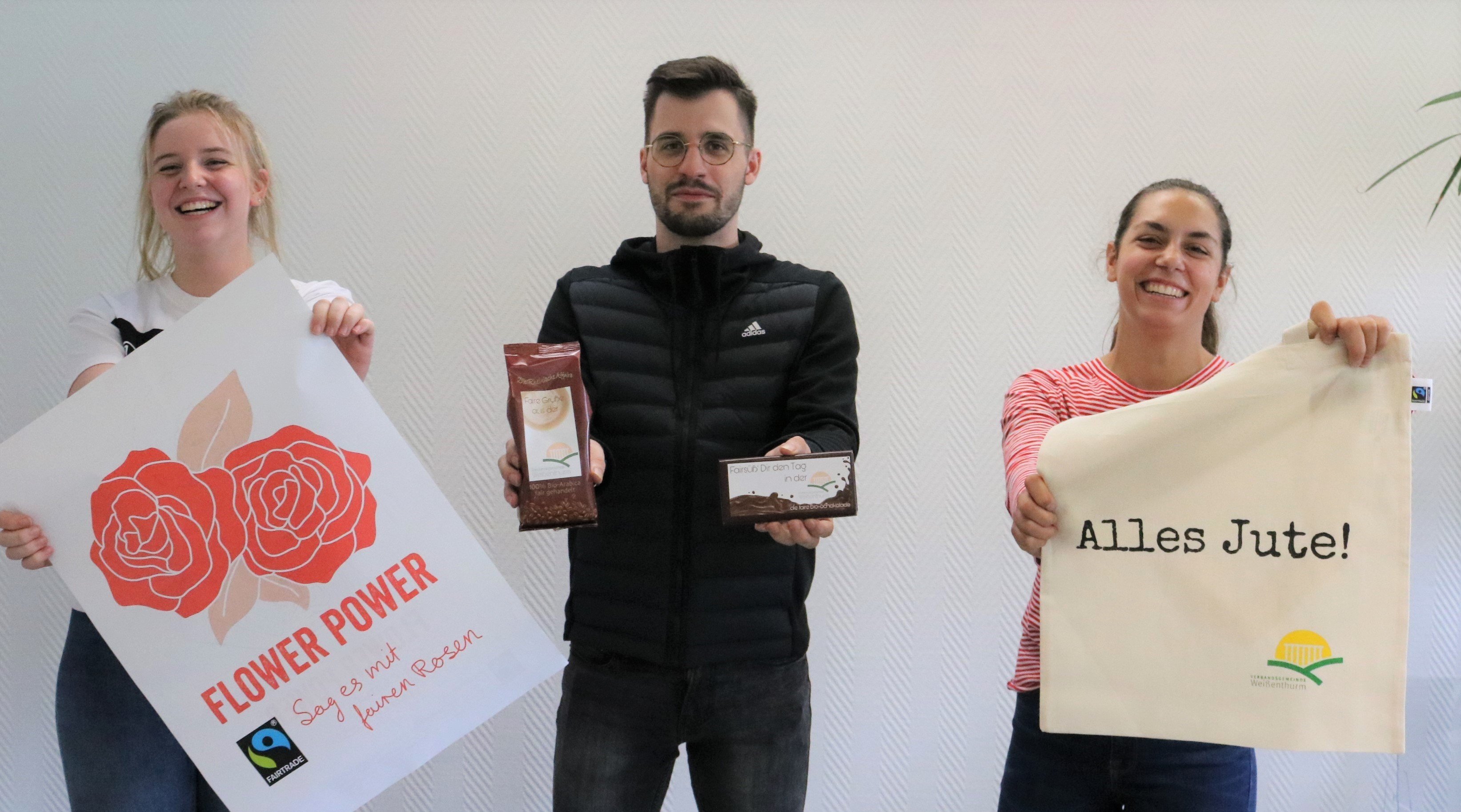 Drei Azubis zeigen ein Plakat zur fairen Woche, Jutebeutel und fair gehandelte Produkte.