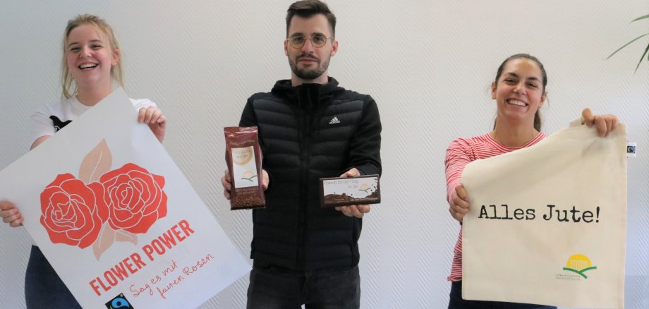Drei Azubis zeigen ein Plakat zur fairen Woche, Jutebeutel und fair gehandelte Produkte.