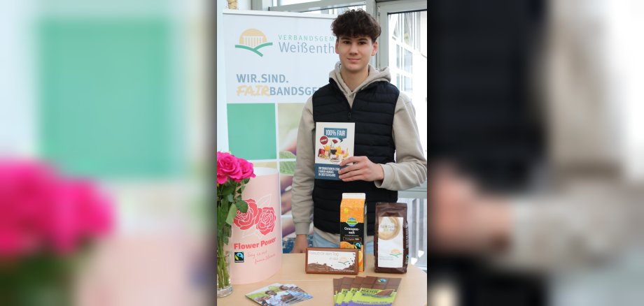 Praktikant Janne Lyding steht im Foyer des VG-Rathauses an einem Stand mit fairen Produkten und Info-Material, in der Hanf hält er eine Broschüre.