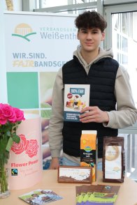 Faire Woche Stand Praktikant Janne Lyding steht im Foyer des VG-Rathauses an einem Stand mit fairen Produkten und Info-Material, in der Hanf hält er eine Broschüre.