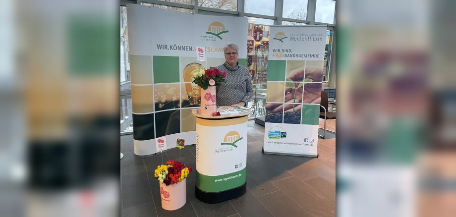 VG-Mitarbeiterin Ulrike Rieden steht an einem Info-Stand mit fairen Rosen und Info-Material im Foyer des VG-Rathauses.