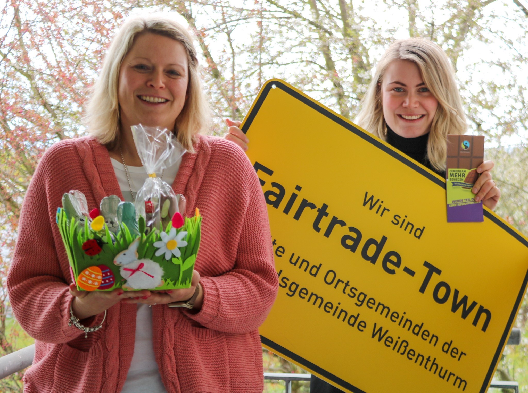 Die VG-Mitarbeiterin Kristina Uerz hat ein Nest mit fairen Produkten in der Hand, neben ihr Kollegin Kim Lachmann mit einem großen, gelben Ortsschild mit der Aufschrift Fairtrade-Town Verbandsgemeinde Weißenthurm. 