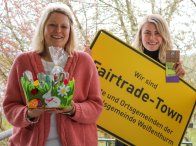 Faire Ostern Die VG-Mitarbeiterin Kristina Uerz hat ein Nest mit fairen Produkten in der Hand, neben ihr Kollegin Kim Lachmann mit einem großen, gelben Ortsschild mit der Aufschrift Fairtrade-Town Verbandsgemeinde Weißenthurm.