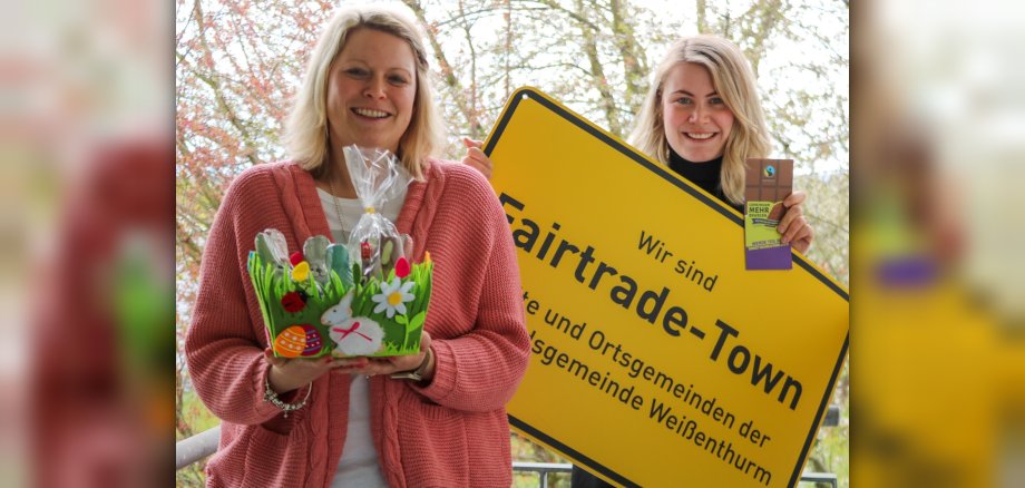 Die VG-Mitarbeiterin Kristina Uerz hat ein Nest mit fairen Produkten in der Hand, neben ihr Kollegin Kim Lachmann mit einem großen, gelben Ortsschild mit der Aufschrift Fairtrade-Town Verbandsgemeinde Weißenthurm. 