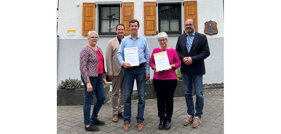 Vor dem Ferienhaus stehen das Gastgeberpaar Gabriele Kohns und Ralf Arnold jeweils mit den Urkunden in einem Bilderrahmen in der Hand, links außen VG-Touristikerin Ulrike Rieden, Bürgermeister Thomas Przybylla daneben und rechts der Kettiger Ortsbürgermeister Peter Moskopp. 