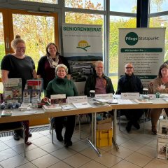 Vor den Rollups des Seniorenbeirates der VG und des Pflegestützpunktes steht die Bassenheimer Bürgermeisterin Natalja Kronenberg ganz links hinter einem Tisch, neben ihr die Gemeindeschwester Daniela Degner. an dem Tisch sitzen die Seniorenbeauftrage Renate Schumann, Seniorensicherheitsberater Norbert Besmens, der Vorsitzende des Seniorenbeirats Hajo Reif, Gabi Schneider vom Pflegestützpunkt und Nina Ketzner vom Bürgerstützpunkt+. 