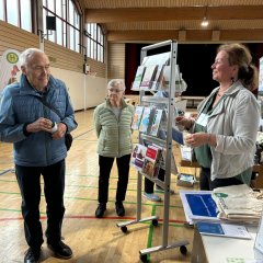 Rechts im Bild steht an einem Infostand mit Ständern für Broschüren die neue Gemeindeschwester Larissa Rieken und informiert einen Senior, der links steht,. Im Hintergrund ist eine ältere Dame zu sehen. 
