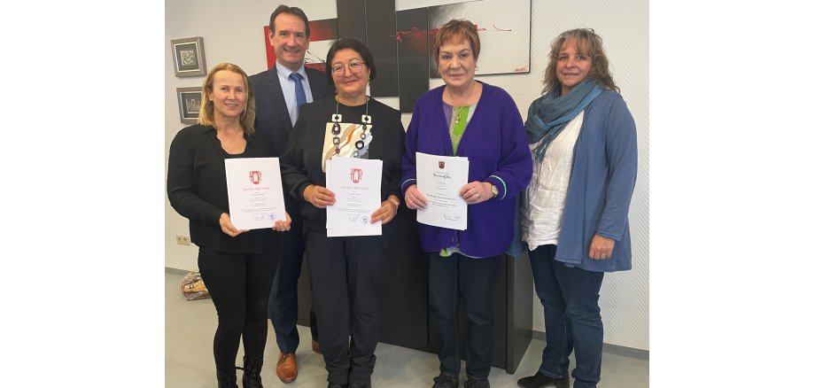 Die drei Jubilarinnen haben ihre Urkunden in der Hand und stehen gemeinsam mit Bürgermeister Thomas Przybylla und Personalratsmitglied Claudia Piske vor einem niedrigen Schränkchen. 