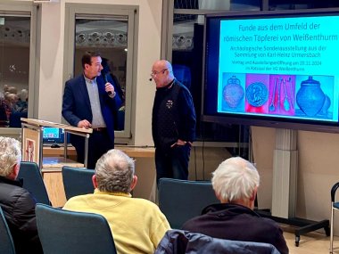 Nahaufnahme von Bürgermeister Przybylla mit Mikrofon am Rednerpult, neben ihm Referent Urmersbach vor dem Bildschirm, auf dem der Titel seiner Präsentation zu sehen ist. Im Vordergrund der Rücken dreier Besucher.