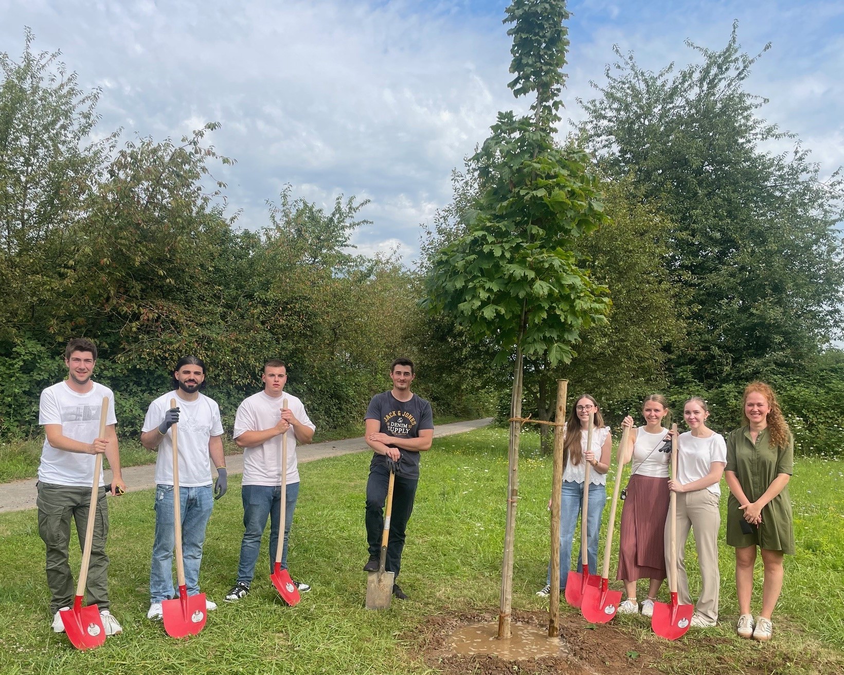 Fünf männliche Azubis stehen mit roten Schaufeln links von dem frisch gepflanzten Ahorn, rechts stehen drei weibliche Azubis, ebenfalls mit roten Schaufeln, und die Ausbildungsleiterin Johanna Püsch. 