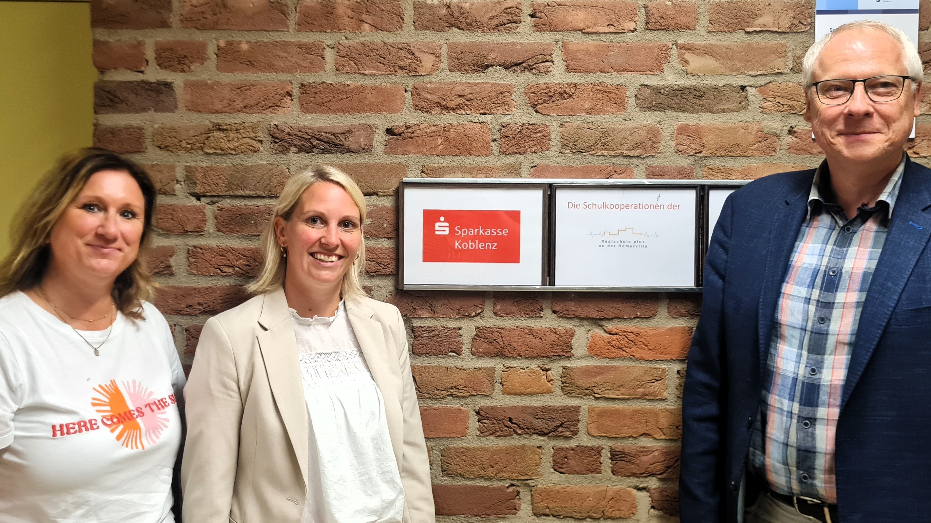 Neben einem Schild mit dem Logo der Sparkasse und der Realschule plus an der Römerville steht rechts Schulleiter Gerhard Müller, links die Ausbildungsleiterin der Sparkasse, Tanja Hommen und ganz links Jobfüxin Katrin Lyding.