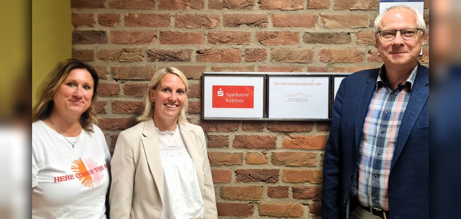 Neben einem Schild mit dem Logo der Sparkasse und der Realschule plus an der Römerville steht rechts Schulleiter Gerhard Müller, links die Ausbildungsleiterin der Sparkasse, Tanja Hommen und ganz links Jobfüxin Katrin Lyding.