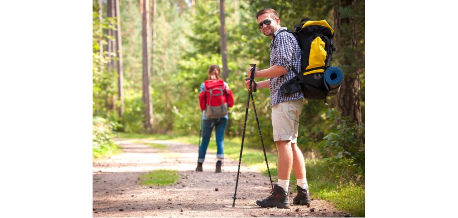 Auf einem Waldweg steht ein jüngerer Mann mit Bermudas, kariertem Hemd und einem schwarz-gelben Rucksack, zwei Wanderstöcke in der Hand, der sich leicht Richtung Betrachter dreht. Weiter vorne auf dem Weg ist eine junge Frau mit einem roten Rucksack von hinten zu erkennen.