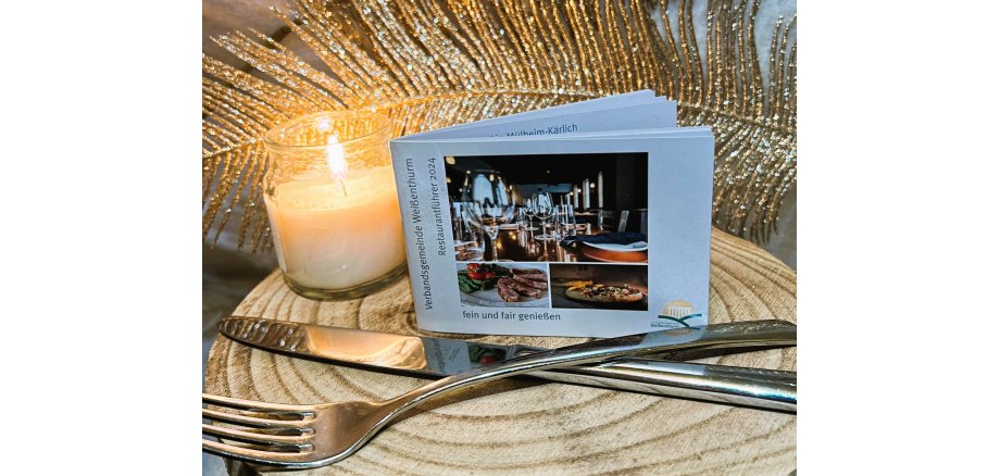 Der Restaurantführer, ein kleines, handliches Heft, steht aufrecht auf einer Holzscheibe neben einer brennenden Kerze, dahinter eine große Feder als Deko