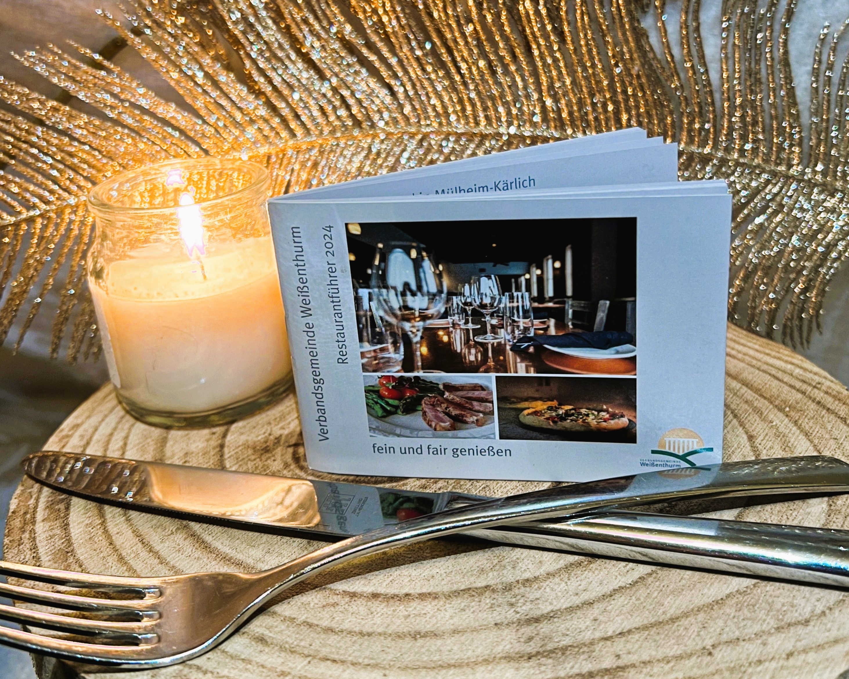 Der Restaurantführer, ein kleines, handliches Heft, steht aufrecht auf einer Holzscheibe neben einer brennenden Kerze, dahinter eine große Feder als Deko