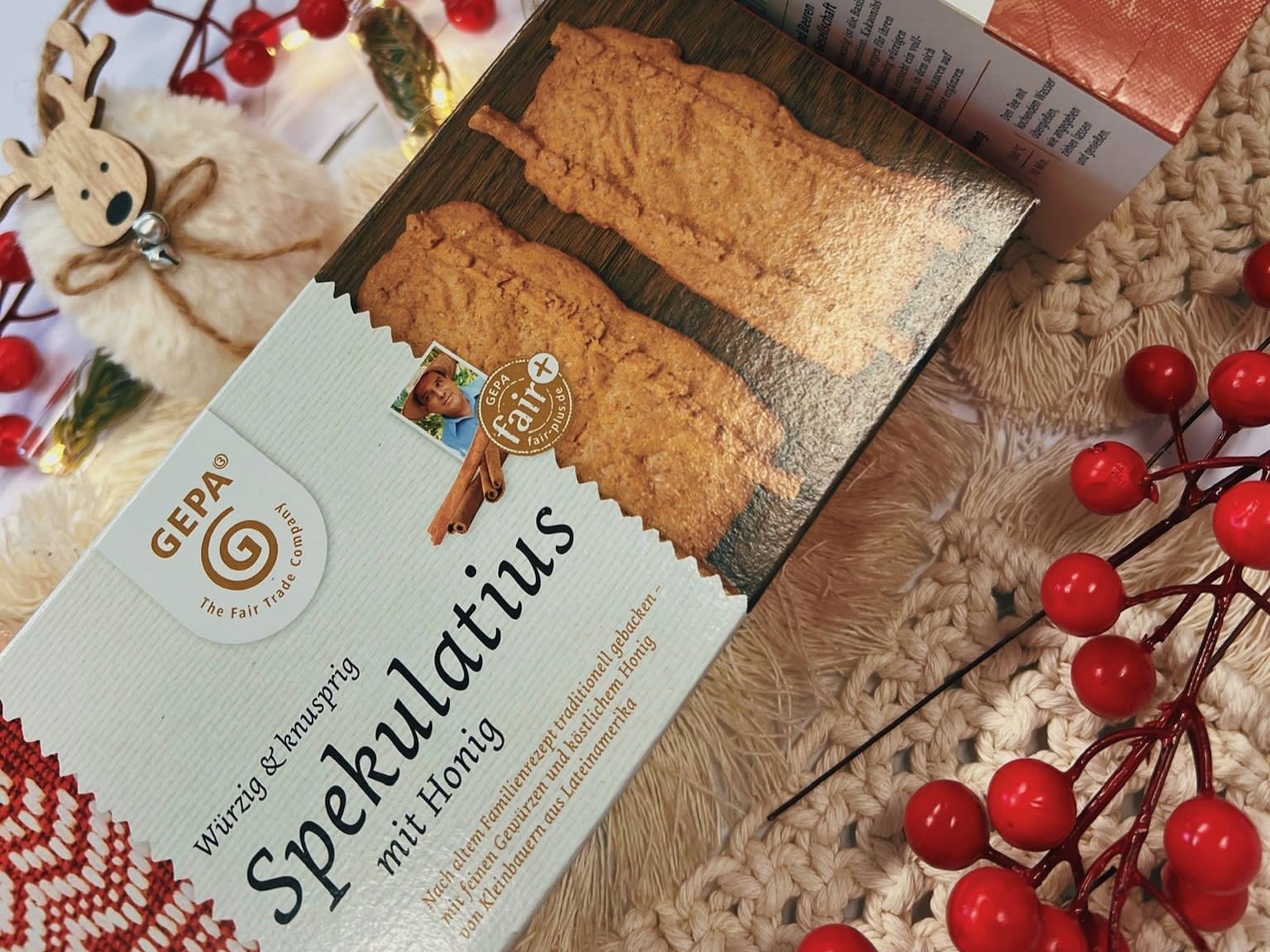 Von links unten nach rechts oben liegt schräg eine Packung Spekulatius auf einem beigen Textiluntergrund. In der linken oberen Ecke ist ein kleiner Plüschelch mit Holzgesicht und -geweih zu sehen, in der rechten unteren Ecke ist ein Zweig mit roten Beeren. 