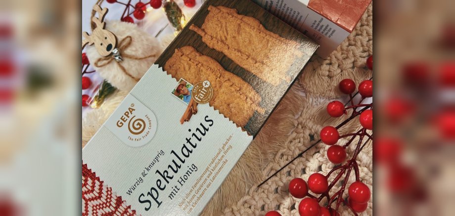 Von links unten nach rechts oben liegt schräg eine Packung Spekulatius auf einem beigen Textiluntergrund. In der linken oberen Ecke ist ein kleiner Plüschelch mit Holzgesicht und -geweih zu sehen, in der rechten unteren Ecke ist ein Zweig mit roten Beeren. 