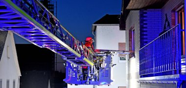 Am linken Bildrand ist eine Drehleiter zu sehen, rechts ist ein Balkon. Die Szene ist in der Dämmerung am frühen Abend.