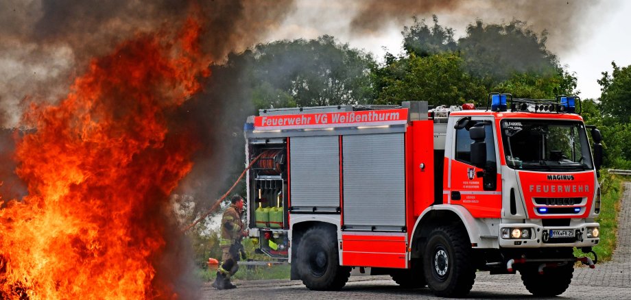 Im linken Vordergrund lodert ein Feuer, rechts weiter hinten steht ein Feuerwehrfahrzeug mit dem Schriftzug Verbandsgemeinde Weißenthurm.