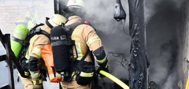 Zwei Feuerwehrmänner sind von hinten zu sehen, wie sie mit einem Schlauch in der Hand einen Brand bekämpfen. Rechts am Bildrand sind verkohlte schwarze Reste und eine unverputzte Wand zu erkennen.