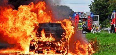 Ein Auto steht in heftigen Flammen im Vordergrund auf einer Straße , dahinter ist ein geöffnetes Feuerwehrfahrzeug mit zwei Feuerwehrleuten zu sehen.