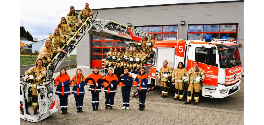 Vor einer Feuerwache steht ein Feuerwehrfahrzeug mit Drehleiter. Die Leiter ist so ausgefahren, dass sie zunächst nach links zeigt und dann bis auf den Boden führt. Darauf stehen Feuerwehrfrauen aus den VG-Einheiten, außerdem direkt vor dem Fahrzeug. Die Mädchen der Jugendfeuerwehr stehen neben dem Korb der Feuerleiter auf dem Boden und bilden eine Abschlussriehe bis zum Fahrzeug. 