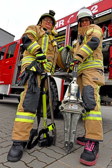 Die Feuerwehrfrauen Miriam Klaß und Patricia Höfer stehen in voller Montur vor einem Fahrzeug, zwischen sich Gerätschaften. Das Foto ist von schräg unten aufgenommen. 