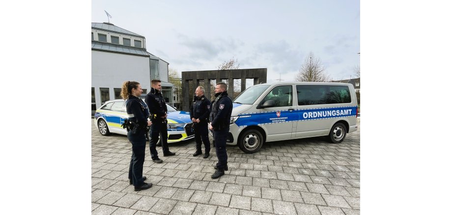Vor dem Rathausgebäude und sieben Säulen stehen links ein Polizeiauto und rechts ein Transporter des Ordnungsamtes so schräg, dass sie sich an den Frontkühlern fast berühren. Davor stehen links eine Polizistin und ein Polizist, rechts zwei Mitarbeiter des kommunalen Vollzugsdienstes. 