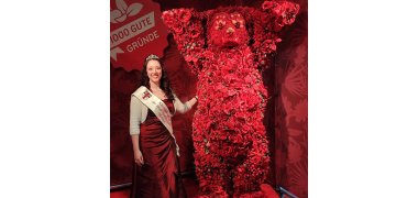 Kirschblütenkönigin Marina I in Kleid und Schärpe steht neben einem roten Berliner Bären mit erhobenen Händen, der aus Früchten und Blumen gestaltet ist
