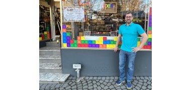 Marc Harrichhausen steht an dem mit bunten Klemmbaustein-Aufklebern beklebten Schaufenster seines Ladens auf der Straße. 