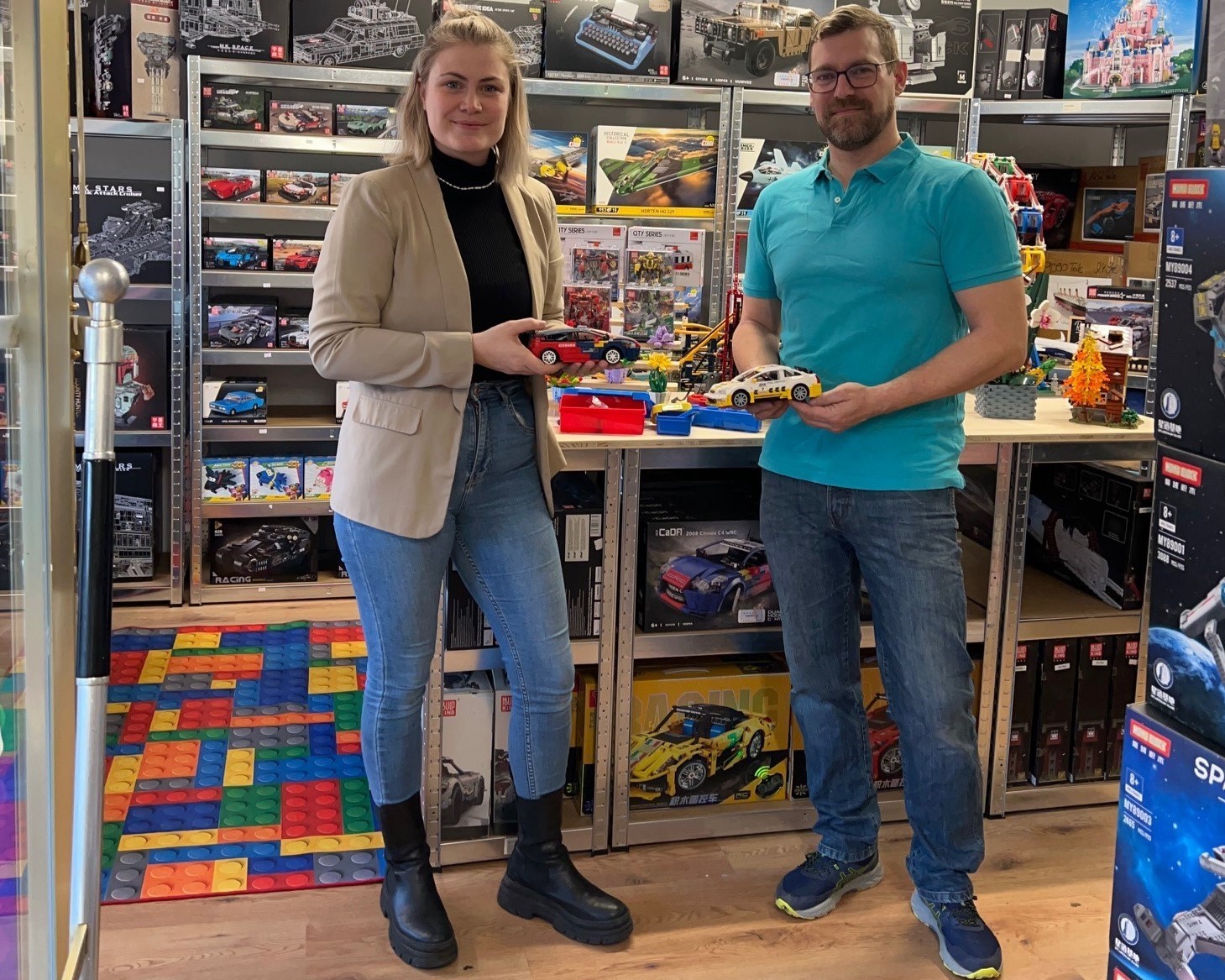 Wirtschaftsförderin Kim Lachmann steht mit Marc Harrichhausen im Laden vor einem Regal mit vielen Packungen. Jeder hält ein Rennauto aus Klemmbausteinen in der Hand. 