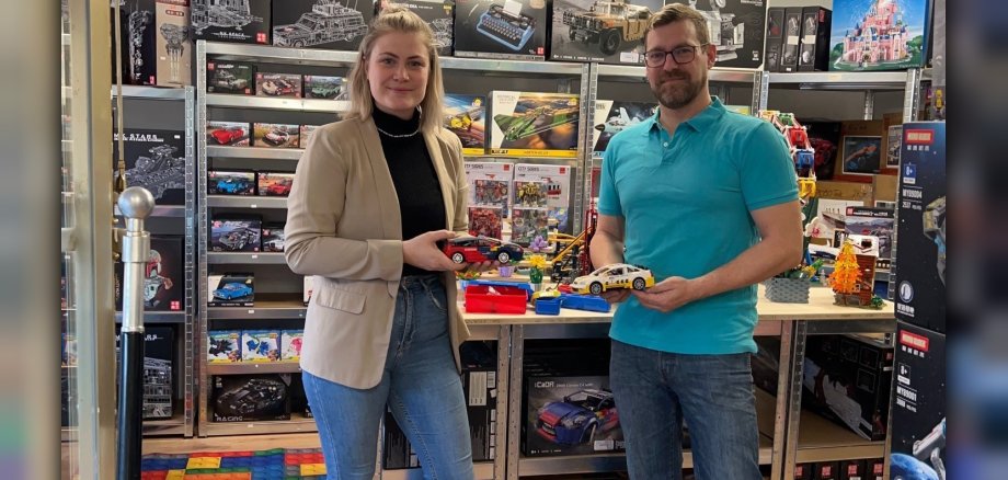 Wirtschaftsförderin Kim Lachmann steht mit Marc Harrichhausen im Laden vor einem Regal mit vielen Packungen. Jeder hält ein Rennauto aus Klemmbausteinen in der Hand. 