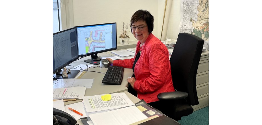 Marita Just trägt eine rote Jacke, sitzt an ihrem Schreibtisch vor der Tastatur mit einer Hand auf der PC-Maus. Beide Bildschirme sind an, vor der Tastatur liegen Mappen und Unterlagen. 