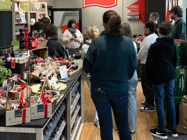 Im Verkaufsraum eines Gewürzherstellers mit einer Theke und Regalen voller Produkte links stehen Schülerinnen und Schüler, die einer Mitarbeiterin zuhören, die links im Hintergrund steht.