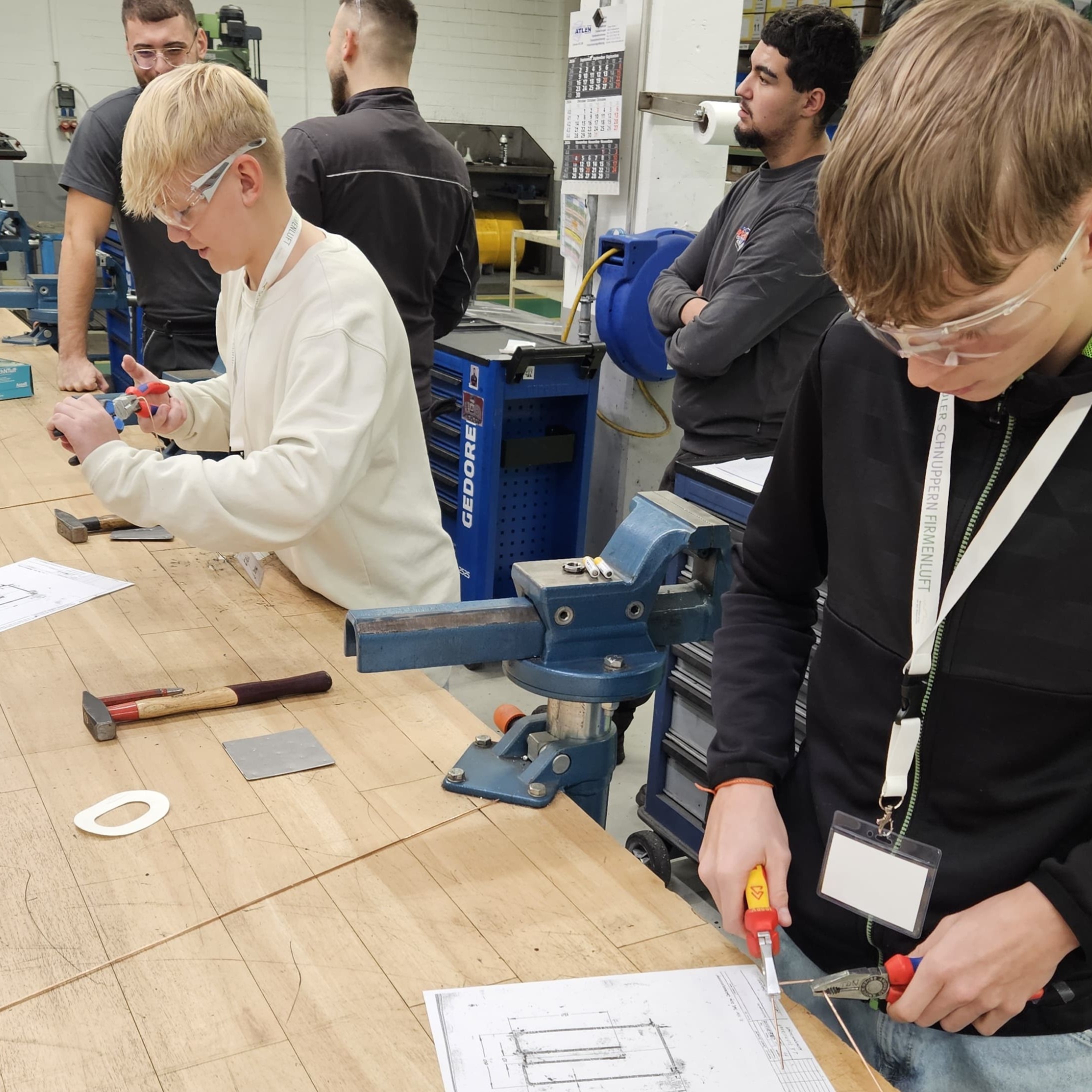 An einer Werkbank stehen stehen zwei Schüler, die Schutzbrillen tragen, und hantieren mit Zangen. Vor ihnen liegt jeweils ein Plan und weiteres Werkzeug, hinter ihnen sind Mitarbeiter des Betriebs und kontrollieren, was die beiden tun.