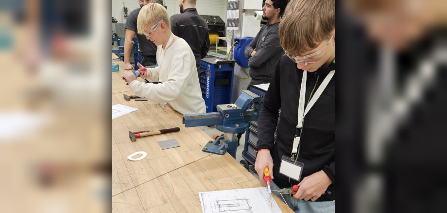 An einer Werkbank stehen stehen zwei Schüler, die Schutzbrillen tragen, und hantieren mit Zangen. Vor ihnen liegt jeweils ein Plan und weiteres Werkzeug, hinter ihnen sind Mitarbeiter des Betriebs und kontrollieren, was die beiden tun.
