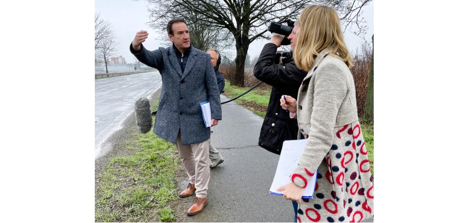 Bürgermeister Thomas Przybylla steht am Straßenrand und gestikuliert, schräg rechts vor ihm ist der Kameramann und die Redakteurin. 