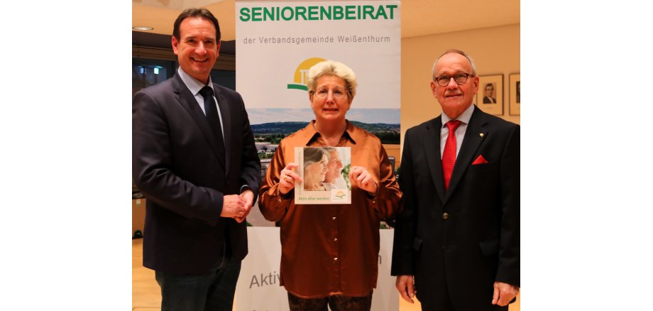 Vor dem Banner des Seniorenbeirats stehen links Bürgermeister Thomas Przybylla, rechts der Vorsitzende des Seniorenbeirats Hajo Reif und in der Mitte VG-Mitarbeiterin Renate Schumann mit der neuen Broschüre für Ältere in der Hand. 