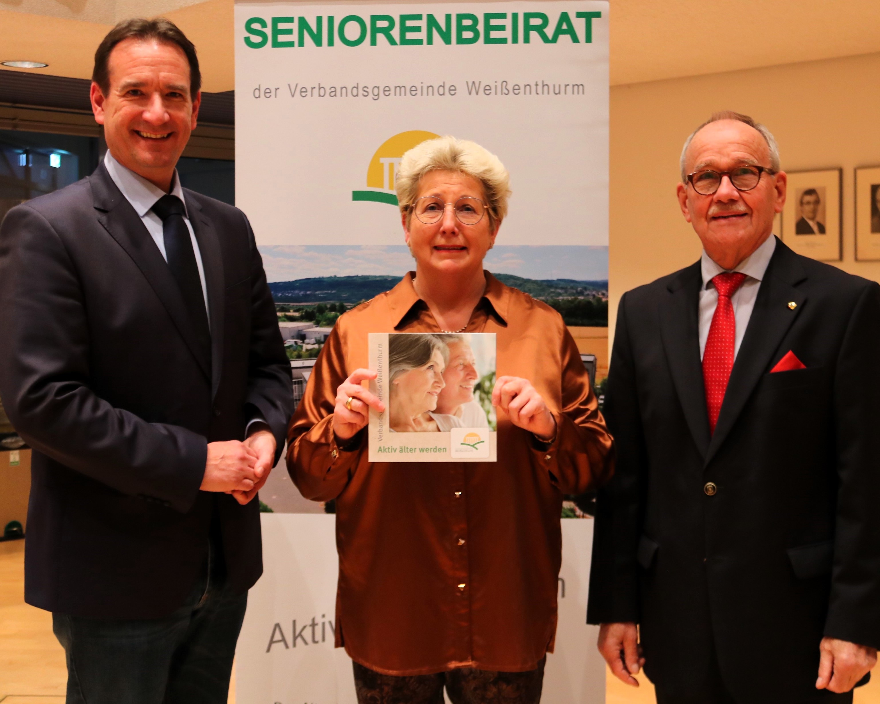 Vor dem Banner des Seniorenbeirats stehen links Bürgermeister Thomas Przybylla, rechts der Vorsitzende des Seniorenbeirats Hajo Reif und in der Mitte VG-Mitarbeiterin Renate Schumann mit der neuen Broschüre für Ältere in der Hand. 