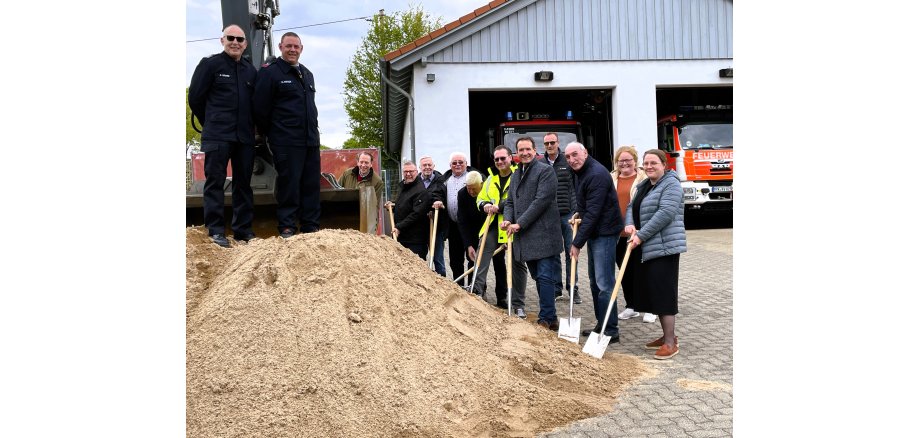 Vor dem Feuerwehrgerätehaus mit geöffneten Toren, hinter denen Fahrzeuge stehen, ist ein Sandhaufen aufgeschüttet, an dem die Teilnehmerinnen und Teilnehmer mit Schaufeln stehen. Auf dem kleinen Hügel stehen die Verantwortlichen der Feuerwehr. 