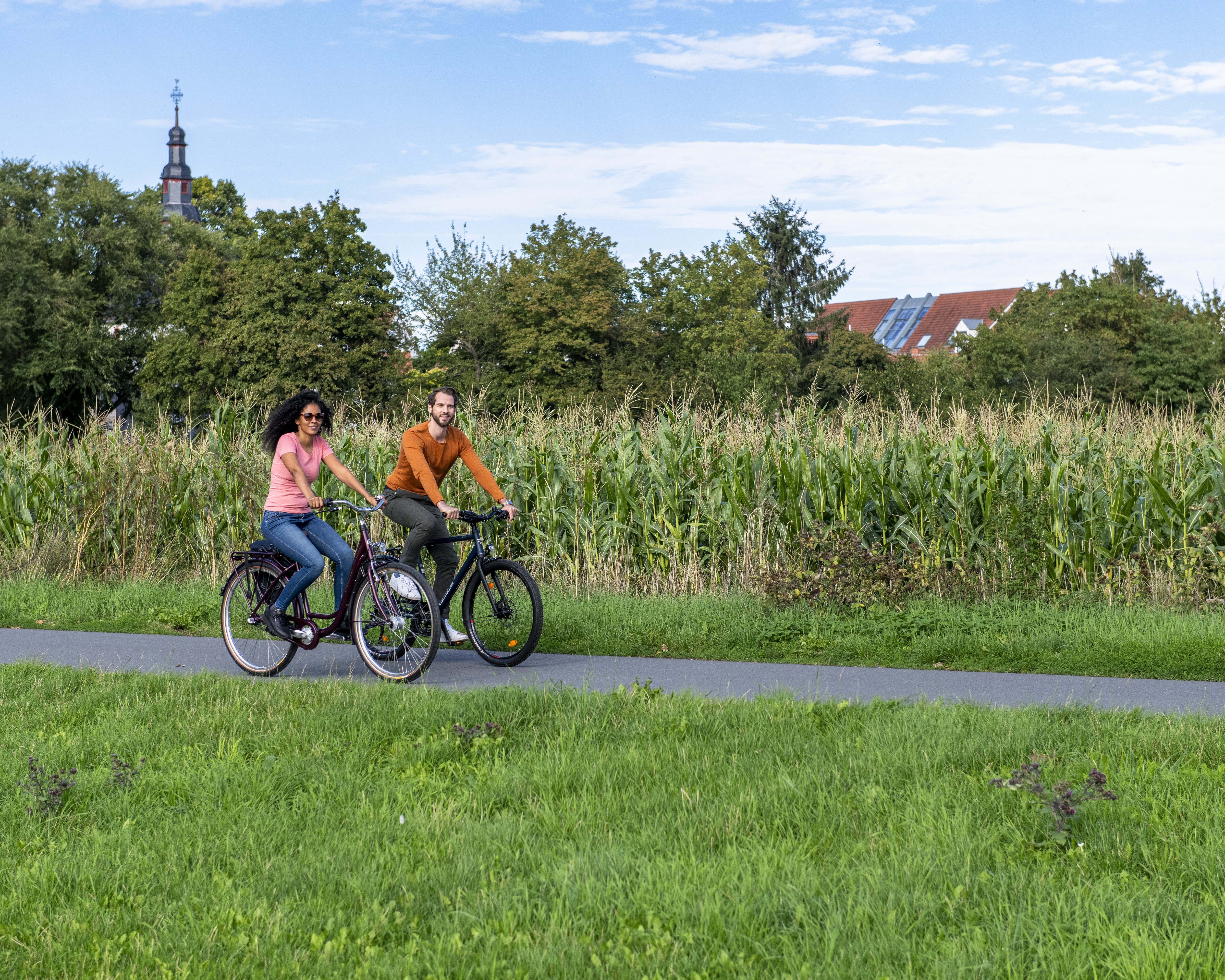 Zwischen einer Wiese und einem Maisfeld fahren von links nach rechts auf Fahrrädern. Die Frau trägt Jeans, ein rosa T-shirt und eine Sonnenbrille und hat langes, schwarzes Haar. Der Mann mit Kinnbart trägt ein braunes Oberteil und eine graue Hose. Im Hintergrund links ragt eine Kirchturmspitze hinter Bäumen heraus, rechts ein Hausdach mit Photovoltaik-Elementen. 