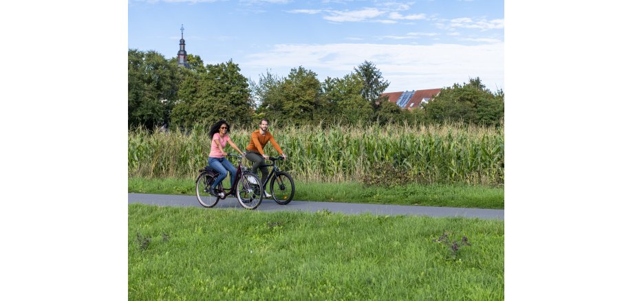 Zwischen einer Wiese und einem Maisfeld fahren von links nach rechts auf Fahrrädern. Die Frau trägt Jeans, ein rosa T-shirt und eine Sonnenbrille und hat langes, schwarzes Haar. Der Mann mit Kinnbart trägt ein braunes Oberteil und eine graue Hose. Im Hintergrund links ragt eine Kirchturmspitze hinter Bäumen heraus, rechts ein Hausdach mit Photovoltaik-Elementen. 