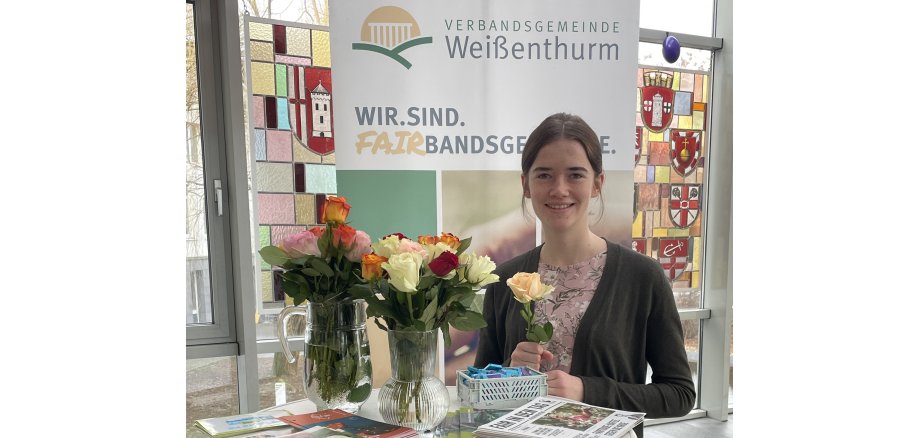 Azubi Sarah Schiller steht an einem Hochtisch mit zwei Vasen voller Rosen und Info-Material. Sie hält eine Rose in der Hand. Hinter ihr ist ein Plakat zur Fairbandsgemeinde zu sehen. 