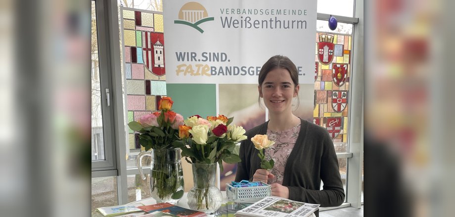 Azubi Sarah Schiller steht an einem Hochtisch mit zwei Vasen voller Rosen und Info-Material. Sie hält eine Rose in der Hand. Hinter ihr ist ein Plakat zur Fairbandsgemeinde zu sehen. 