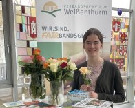 Azubi Sarah Schiller steht an einem Hochtisch mit zwei Vasen voller Rosen und Info-Material. Sie hält eine Rose in der Hand. Hinter ihr ist ein Plakat zur Fairbandsgemeinde zu sehen.