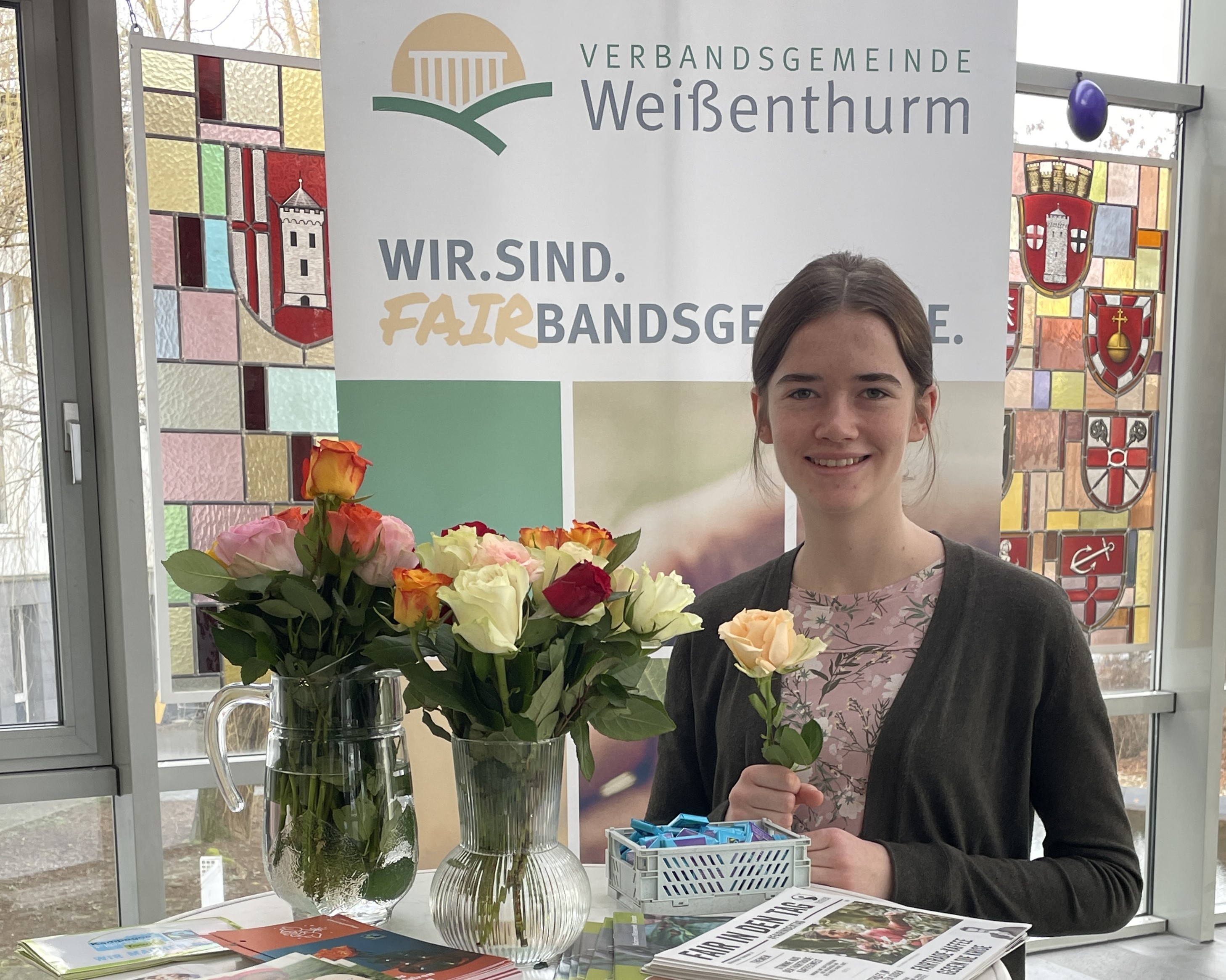 Azubi Sarah Schiller steht an einem Hochtisch mit zwei Vasen voller Rosen und Info-Material. Sie hält eine Rose in der Hand. Hinter ihr ist ein Plakat zur Fairbandsgemeinde zu sehen. 