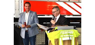 Bürgermeister Thomas Przybylla hält eine Ehrennadel in der Schmuckschachtel in der Hand. Rechts neben ihm steht neben einem Rednerpult Landrat Dr. Alexander Saftig und verliest eine Urkunde. Im Hintergrund ist der Ausschnitt eines Feuerwehrfahrzeugs zu erkennen.