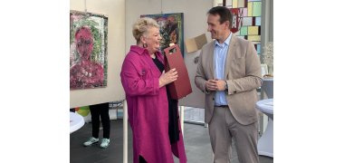 Bürgermeister Thomas Przybylla (rechts) überreicht vor Schautafeln mit Bildern der Künstlerin an Annelie Döpp einen Geschenkkarton mit zwei Saftflaschen darin. 