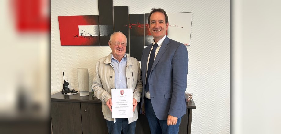 Antonius Rüsing steht links von Bürgermeister Thomas Przybylla vor einem Sideboard und hält seine Ernennungsurkunde in der Hand. 
