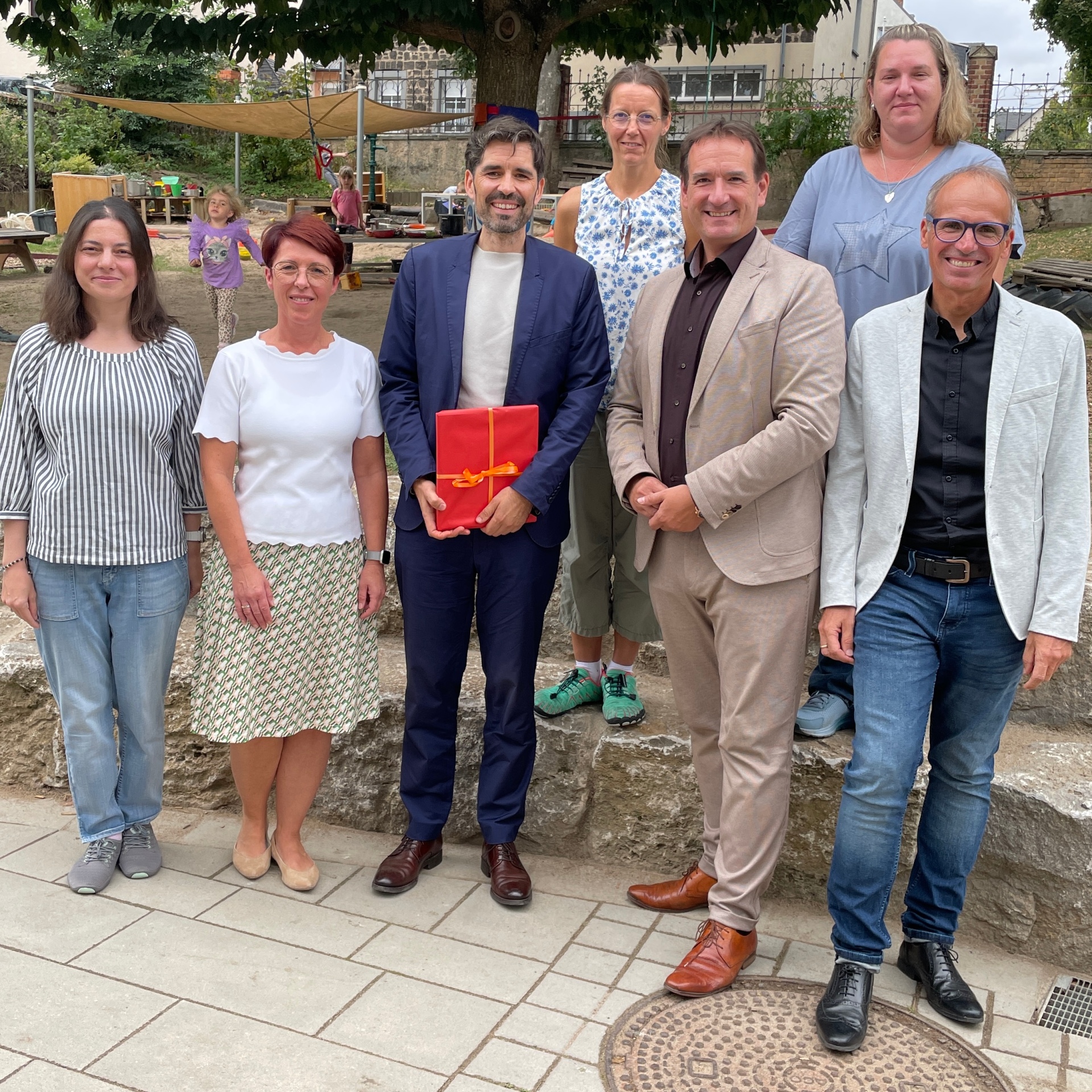 Mit Minister Sven Teuber steht Bürgermeister Thomas Przybylla, Fachbereichsleiter Sven Normann, Kita-Leiterin Tanja Reuter und die drei Kolleginnen des multiprofessionellen Teams im Garten der Kita Bassenheim. 
